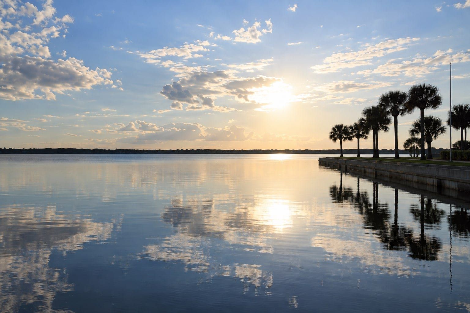 A wide calm lake at sunset under a pale sky with scattered clouds, a low seawall along the right edge with a row of dark palms and a flagpole standing on it